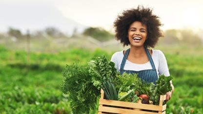 Mujer sonriente en un delantal con una caja de madera de productos frescos saliendo de un campo