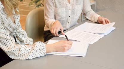 Dos personas revisando y discutiendo documentos en una mesa, una señalando y la otra tomando notas.